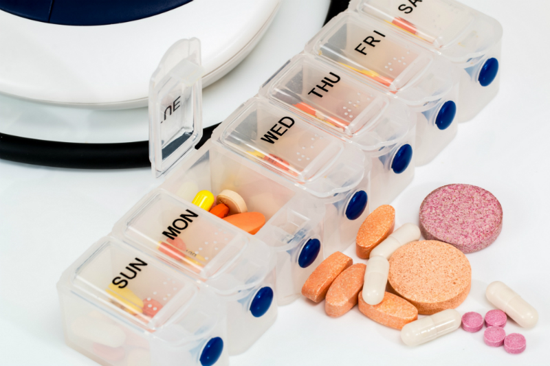 A variety of medications are next to a pill organizer that has one flap up for a particular day’s dose.