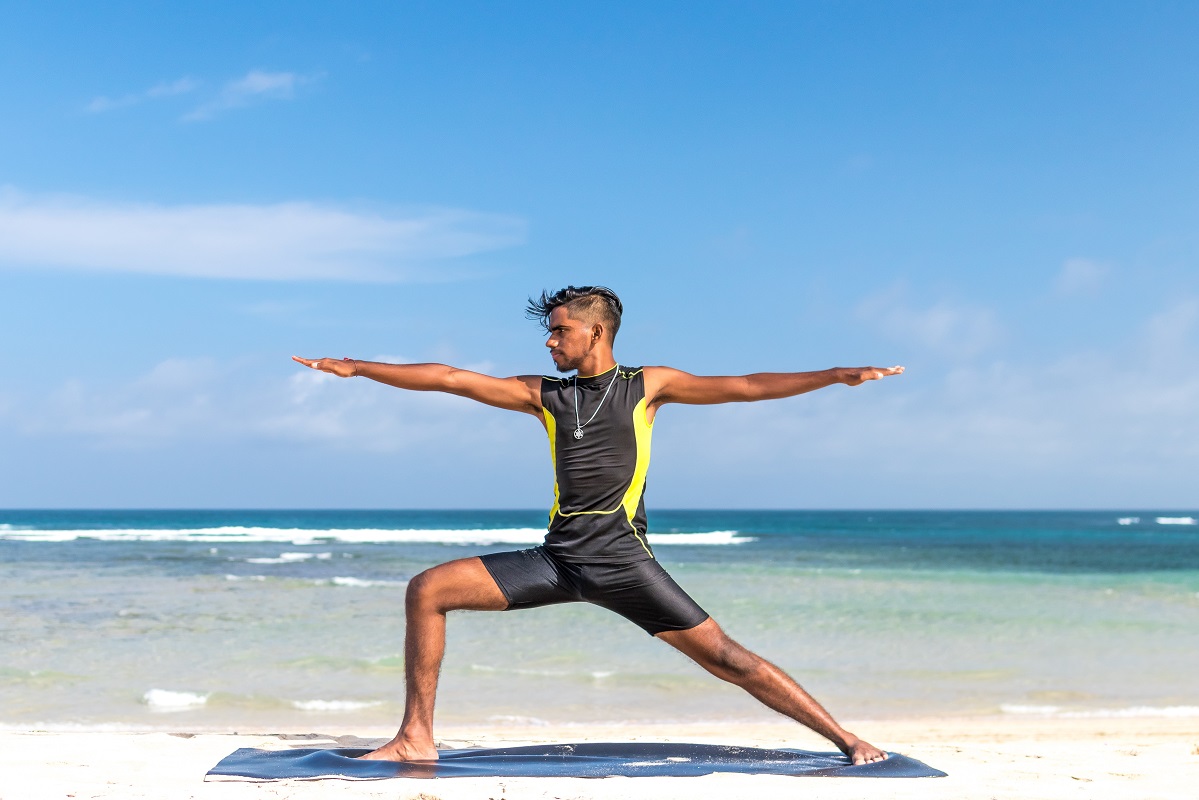 Man in Warrior two pose on yoga mat at beach on sunny day