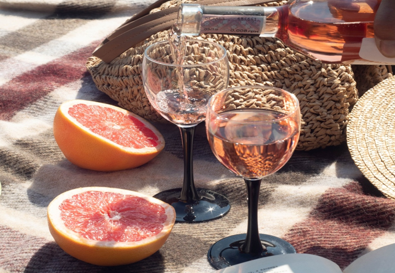 On a picnic blanket, two slices of grapefruit rest near a bag and two glasses of wine; one of which a person is pouring wine into.