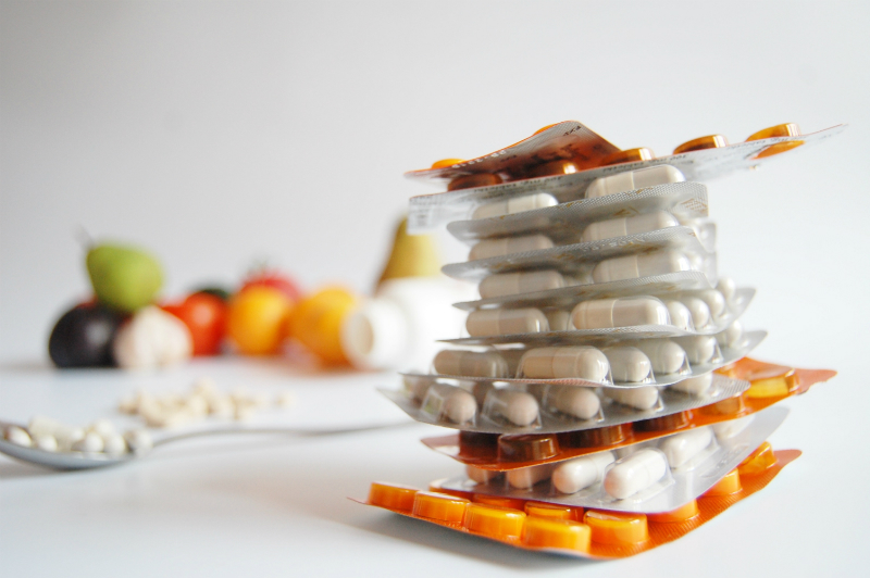 Several fruits rest in the background while packets of pills are stacked on top of one another in the foreground. 