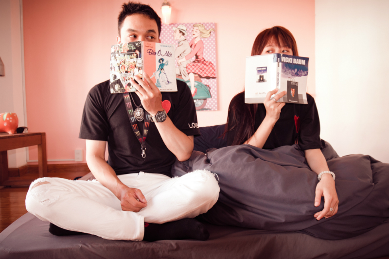 A man and a woman are sitting beside each other on a gray bed and looking over the tops of their books to stare at one another.