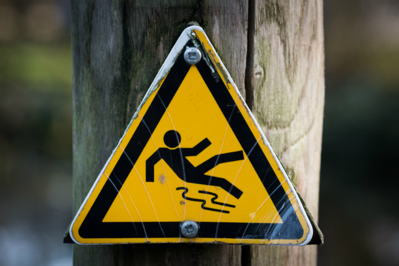 A yellow triangular sign attached to a pole depicts a cartoon man slipping on a road.
